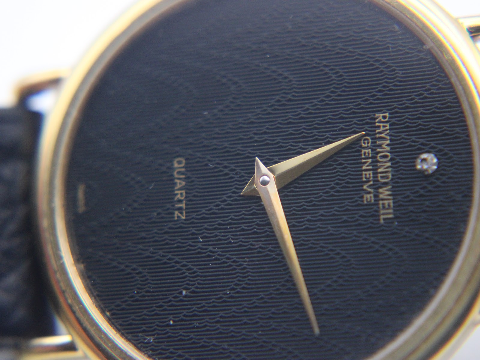 Macro shot of the black textured dial with a subtle wave pattern, gold tone hands, and the single diamond marker at 12 o'clock.