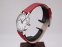 Angled side view of the Mondaine watch showing the silver case and red leather strap, on a watch stand.