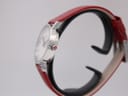 Side view of the Mondaine watch case and red leather strap on a watch stand, highlighting the polished stainless steel finish and signed crown with 'M' logo.