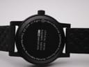 Bottom-up view of the Mondaine watch with its black rubber strap extended, showing the textured underside and case back