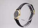 Angled side view of the Raymond Weil watch from the right, showing the gold-tone crown and the profile of the round case and black leather strap.