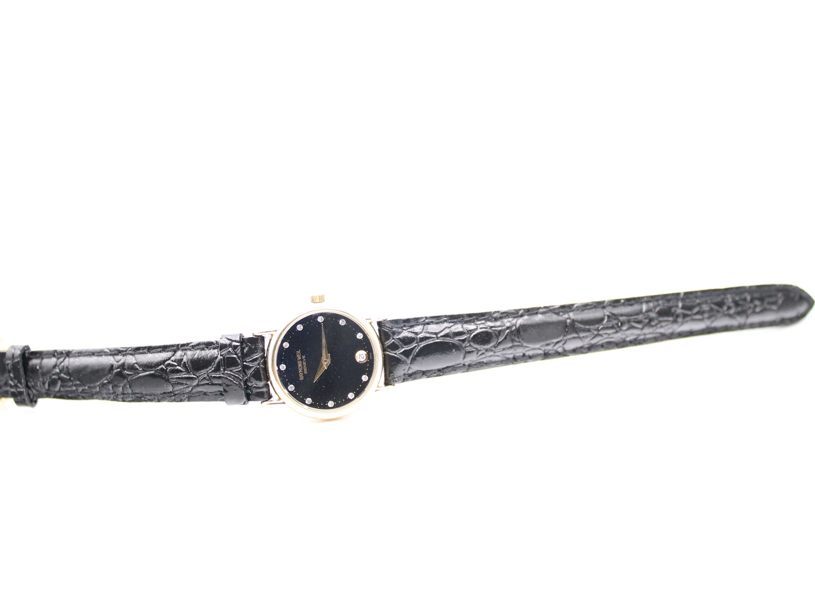 Top-down view of the Raymond Weil watch laid flat, showcasing the full length of the black alligator calf leather strap and the gold-plated watch head.