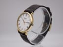 Angled view of the Raymond Weil dress watch on a clear display stand, showing the right side of the gold case and crown.