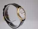 Side view of the Raymond Weil Geneve watch on a clear display stand, showing the gold case and brown leather strap.