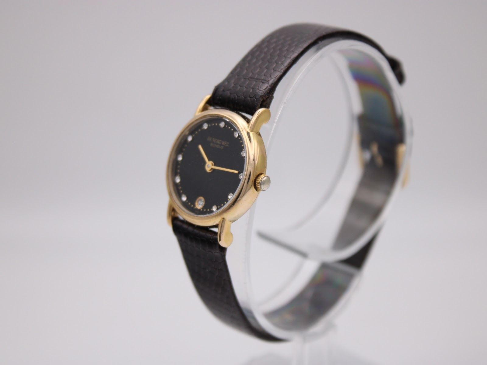 Angled view of the Raymond Weil Geneve watch, showing the gold case, black dial with diamonds, and the texture of the dark brown leather strap.