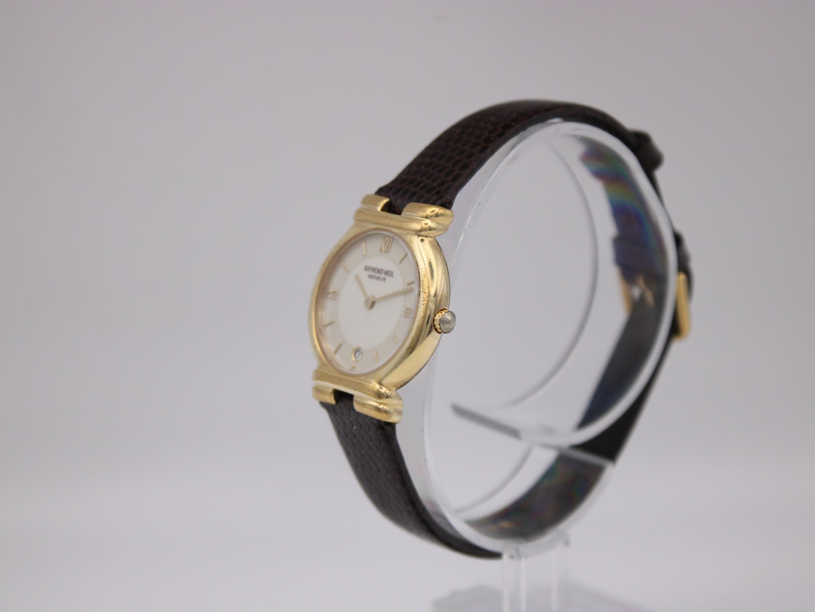 Side view of the Raymond Weil watch, showing the gold-tone case, crown, and curved lugs on a display stand.