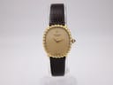 Front view of Raymond Weil 1620 gold tone oval watch on a stand, showing champagne dial with single diamond marker and dark brown strap.