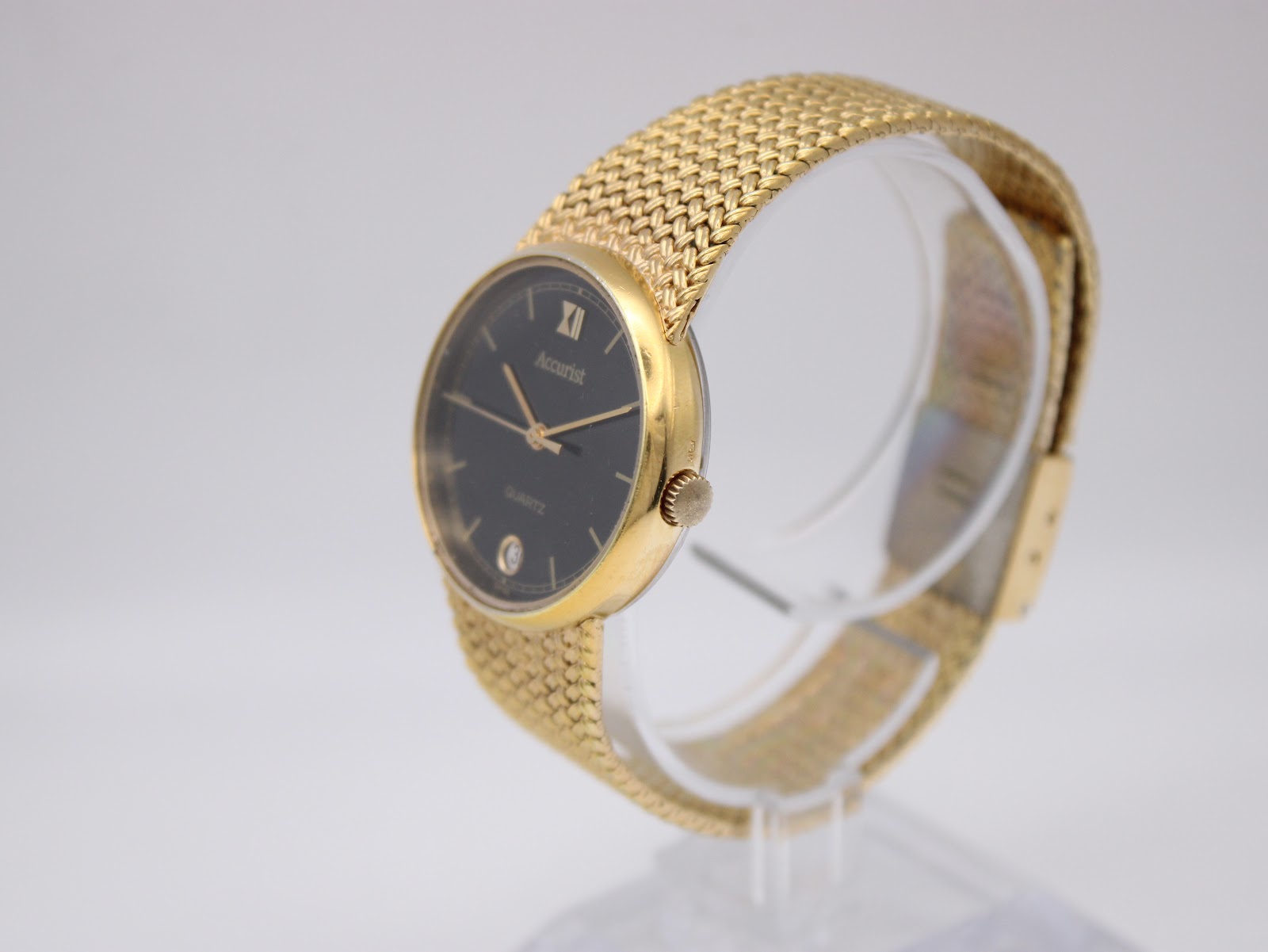 Three-quarter angled view of the Accurist watch on a clear display stand, highlighting the gold case and mesh bracelet.