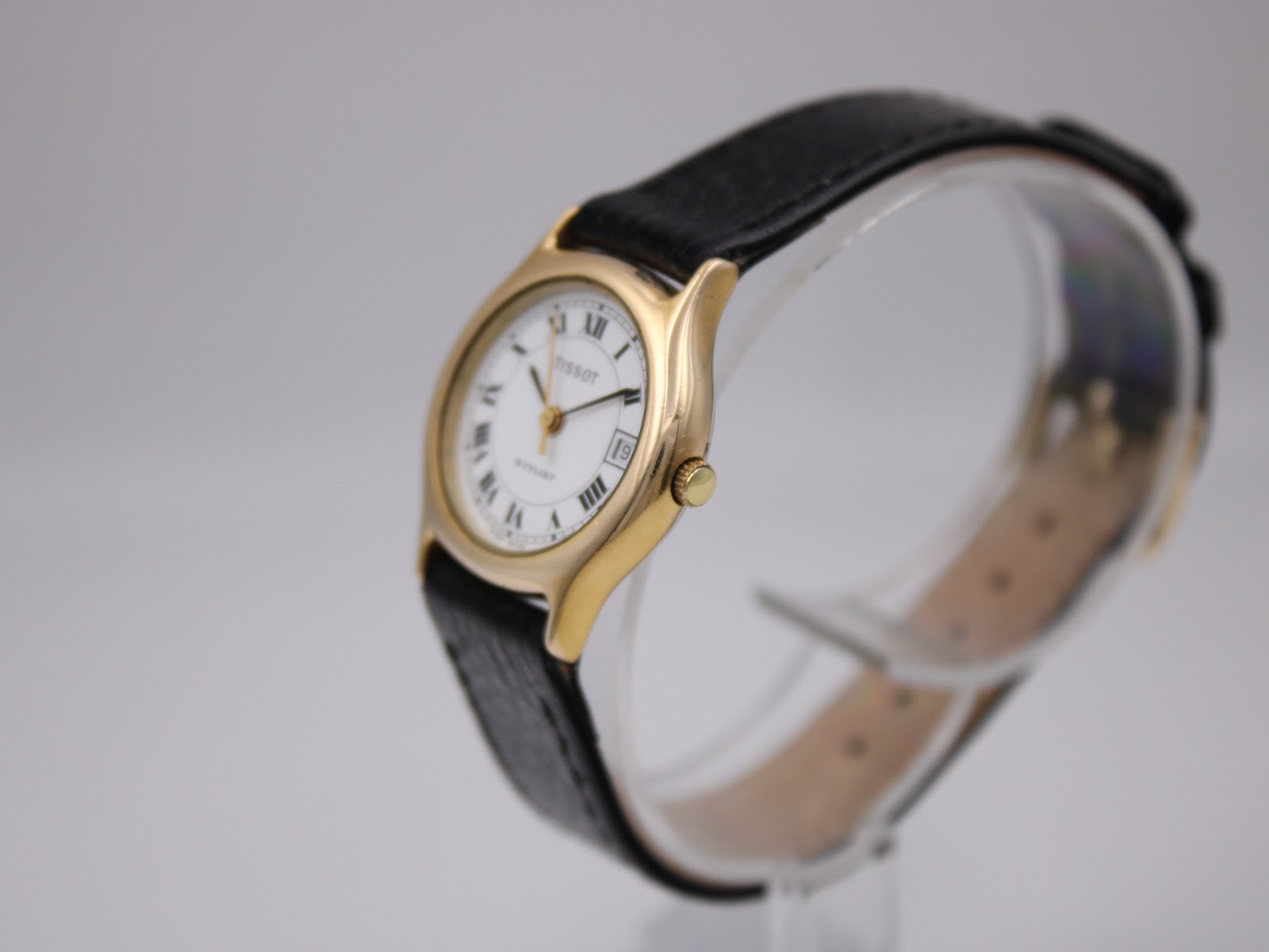 Oblique front-right view of the Tissot Stylist on a clear stand, showing gold case and black leather strap