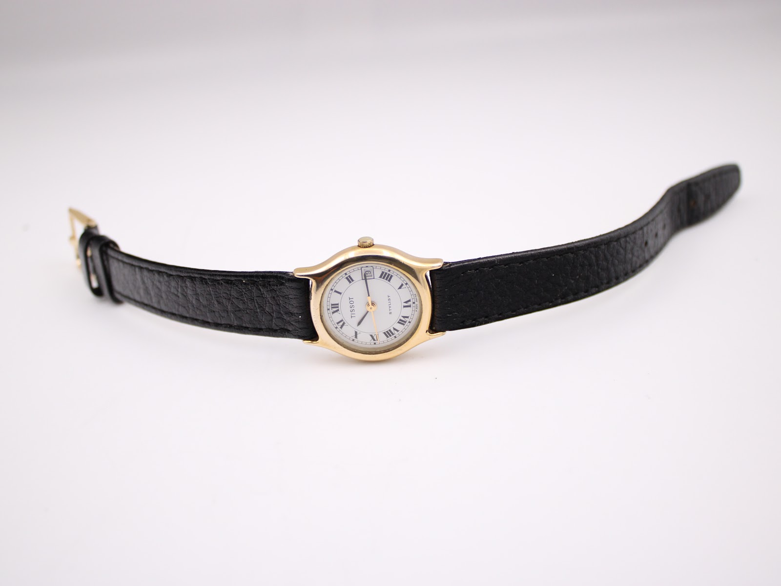Full view of the Tissot Stylist watch laid flat, displaying the gold case and black leather strap with buckle