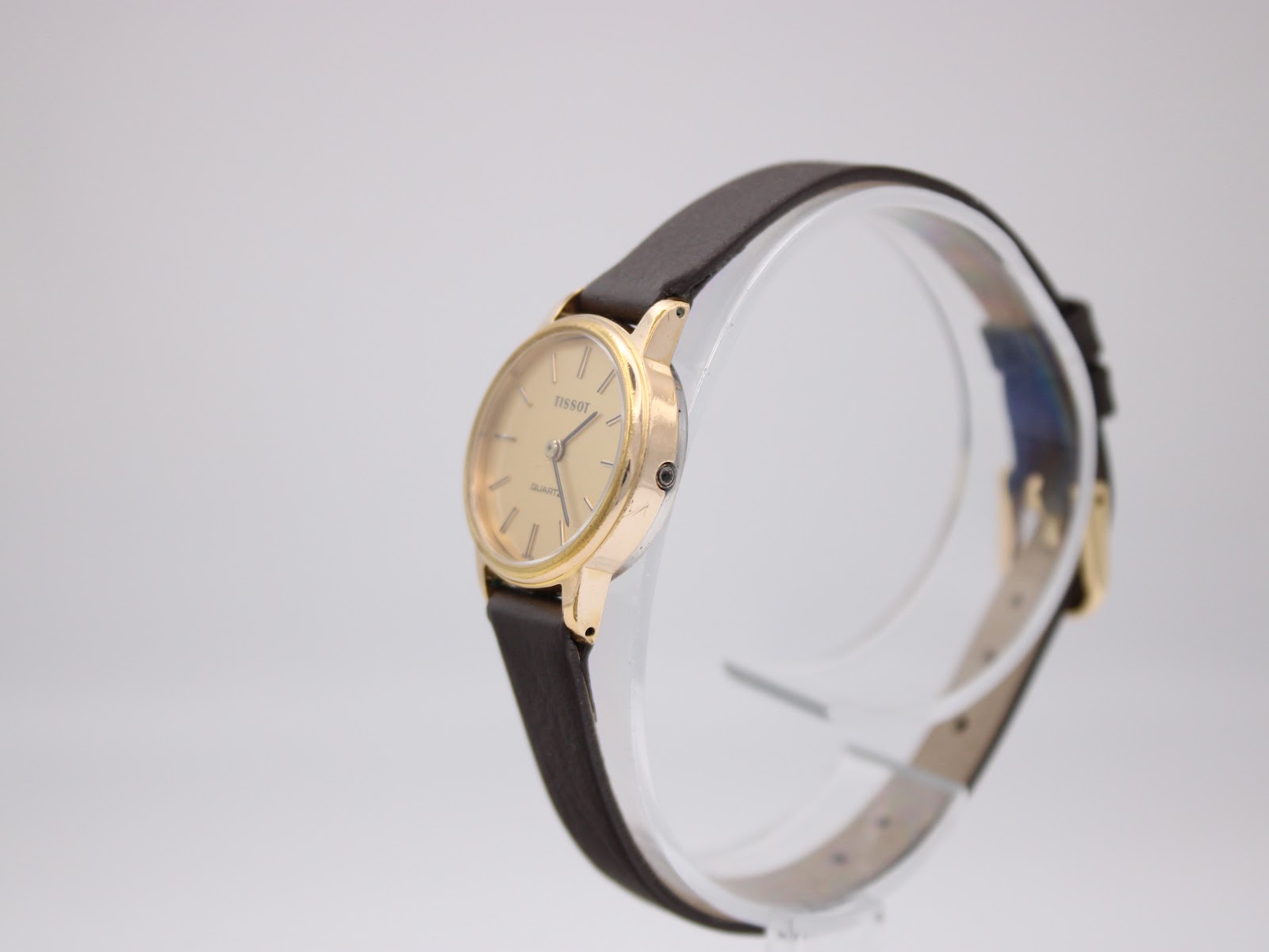 Angled side view of the gold-plated Tissot watch on a clear stand, showing the case profile.