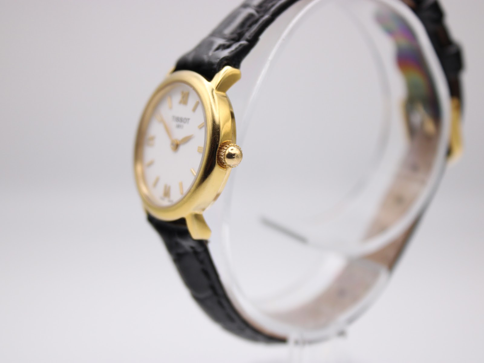 Angled side view of the Tissot C226K watch, showing the gold plated case and crown on a clear stand.