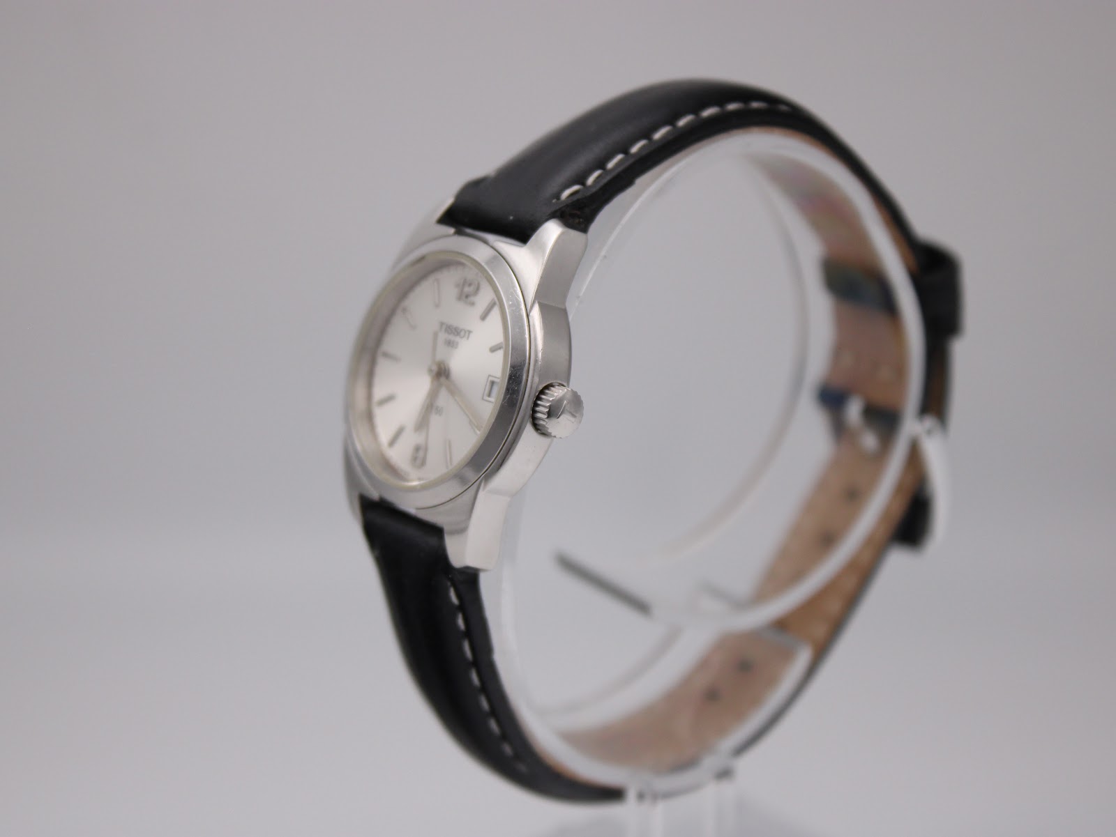 Side view of the Tissot watch on a clear stand, showing the crown and polished stainless steel case.