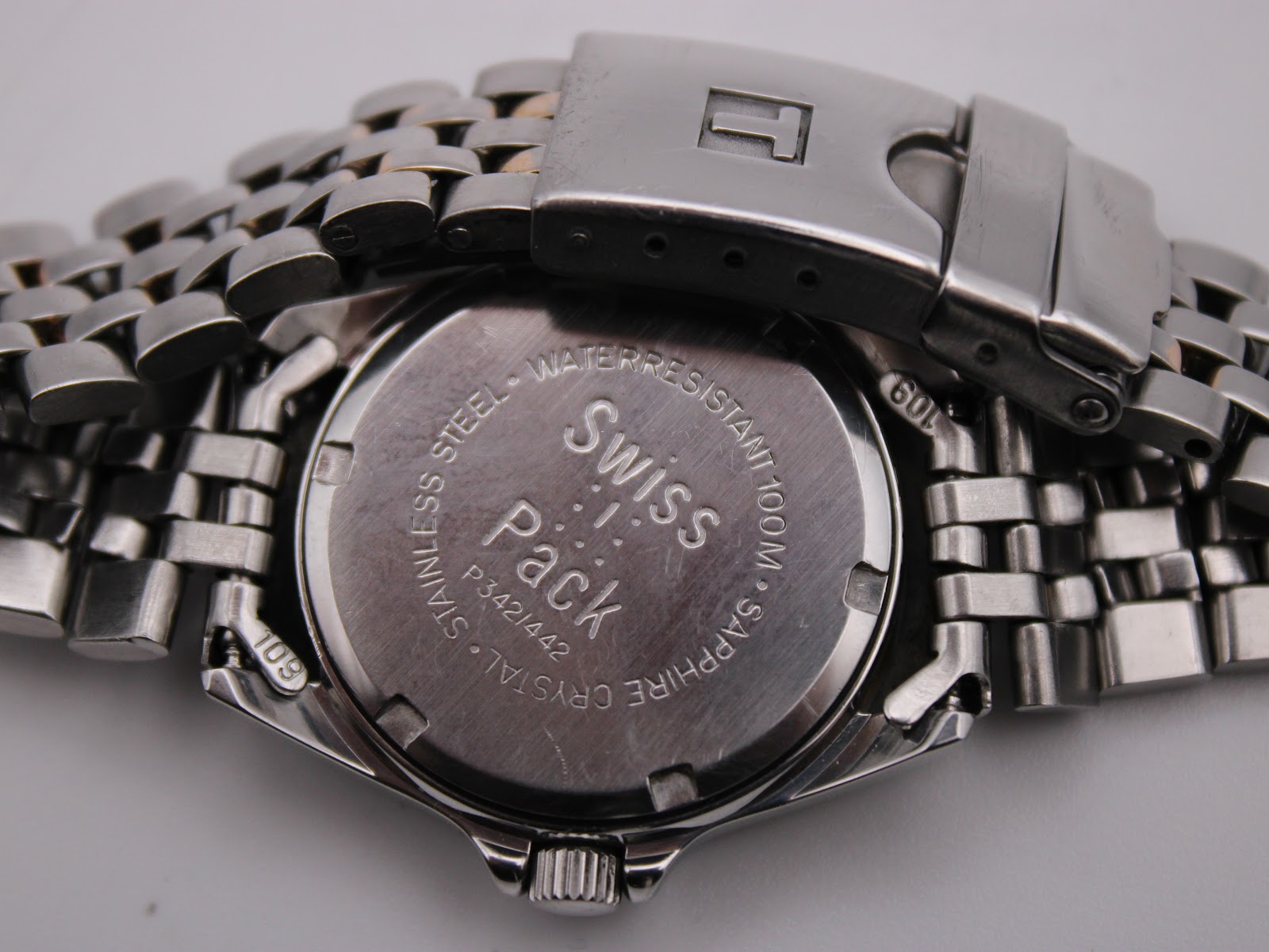 Close-up of the watch's stainless steel case back, clearly showing 'Swiss Pack', 'P342/442', 'Water Resistant 100M', 'Sapphire Crystal' engravings, and the Tissot signed deployant clasp.