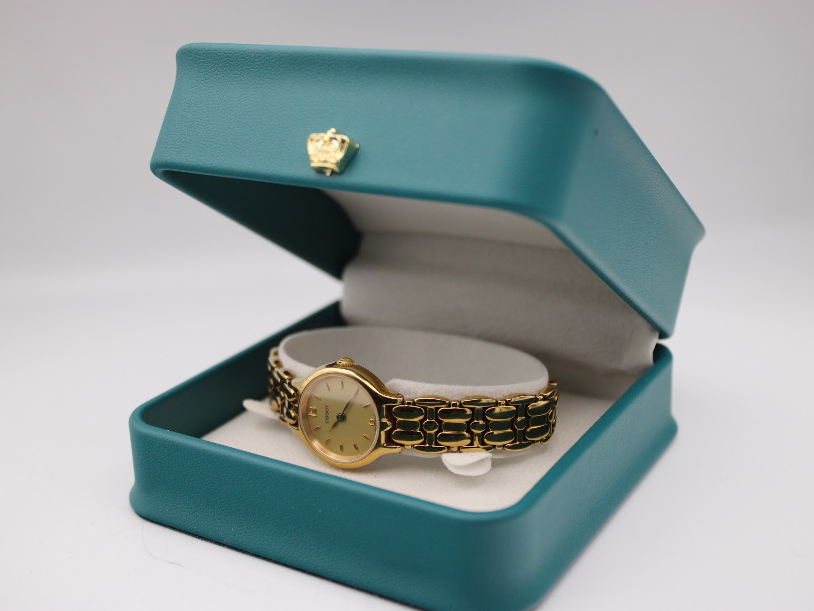 The gold tone Tissot watch displayed inside a decorative teal jewellery box with a gold crown emblem.