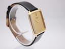 High-angle side view of the gold Raymond Weil watch on a transparent stand, highlighting the rectangular case and black strap.
