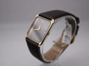 Oblique left side view of the Raymond Weil 9118 on a clear watch stand, showing the rectangular gold case and brown strap.