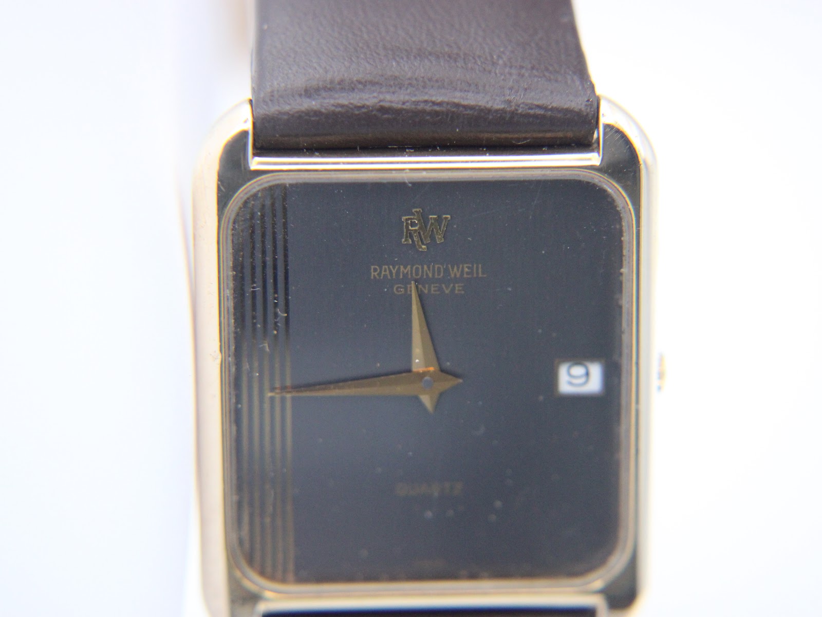 Close-up front view of the Raymond Weil 9118 dial, showing "RW" logo, "Raymond Weil Geneve", "Quartz", gold Dauphine hands, date window, and pinstripe texture.