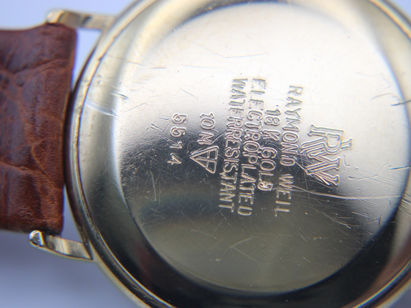 Close-up of the watch case back showing "Raymond Weil Geneve", "18 K Gold Electroplated 10 M", "Water Resistant", and model number "5514" with surface scratches.