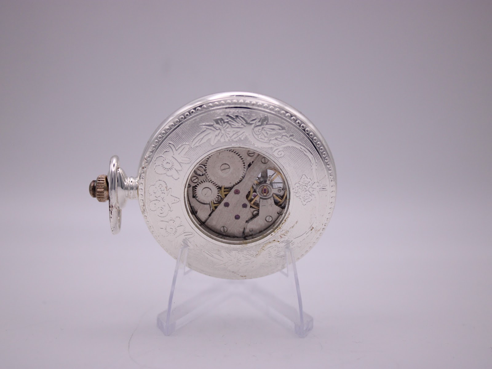 Back view of The Heritage Collection pocket watch on a stand, showing the exhibition case back revealing the manual wind movement and engraved detailing.